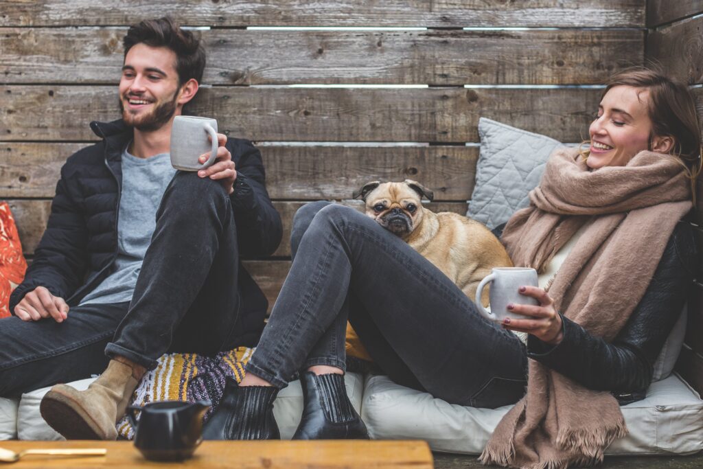 Paar sitzt gemütlich bei einem Kaffe in einer Kuschelecke aus Holz und lächelt. Sich in der Beziehung gelassen zu begegnen ermöglicht so manches Lächeln.